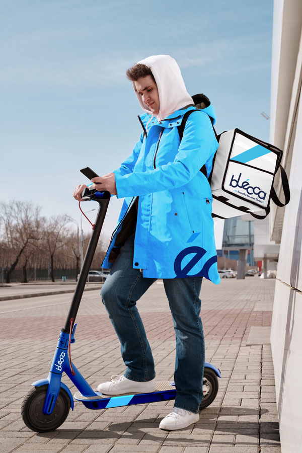 livreur avec une veste bleu clair et le "é" de blécca, il est avec une trottinette bleu au couleur de blécca bleu clair et foncé. Il à un sac pour transporter les commandes de couleurs blanc, avec une bande bleu et le logotype de blécca.